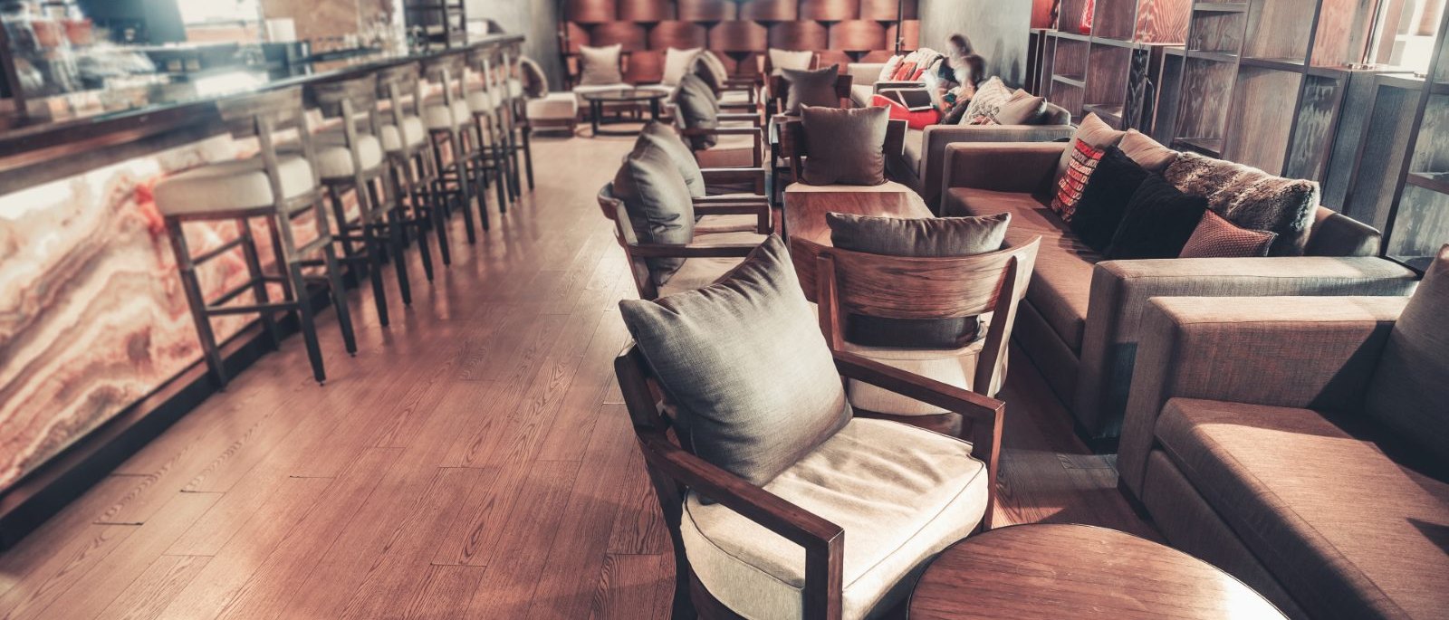 The length of Dionysus in warm light, a lit stone bar and its stools on the left, low armchairs and long sofas grouped along the right, two guests talking at the far end