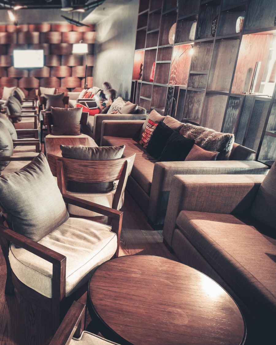 Long sofas with patterned cushions and a fur throw along a wall of lit timber shelving, low armchairs and round tables in front of them